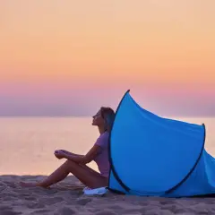 Kobieta siedzi na plaży przy rozłożonym namiocie plażowym. Zachód słońca nad morzem. Jak złożyć namiot plażowy?