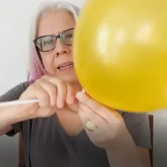 Kobieta w okularach mocuje żółty balon foliowy do białego patyka.