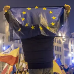 Osoba trzyma flagę UE, symbol jedności, która powstała w 1993 roku. W tle widać inne flagi i tłum ludzi.