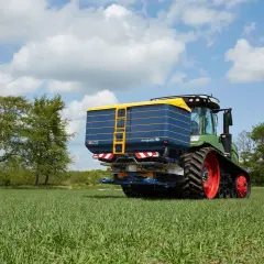 Zielony traktor z przyczepą rozsiewającą na polu. Na przyczepie widoczna nazwa "bogballe tabela wysiewu".