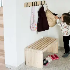 Dziecko wiesza torbę na haczyku. Obok stoi prosta, drewniana ławka, idealna, by szybko zrobić miejsce na buty.