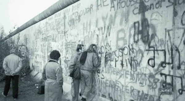 Ludzie oglądają graffiti na Murze Berlińskim, gdy zburzono mur. Widać napisy "Jump over here" i "A house divided cannot stand".