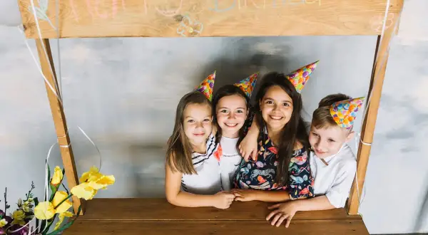 Dzieci w czapkach urodzinowych pozują przy drewnianej konstrukcji z napisem "Happy Birthday". To świetny pomysł, jak zrobić fotobudkę z kartonu na przyjęcie!