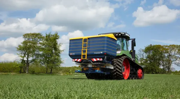 Zielony traktor z przyczepą rozsiewającą na polu. Na przyczepie widoczna nazwa "bogballe tabela wysiewu".