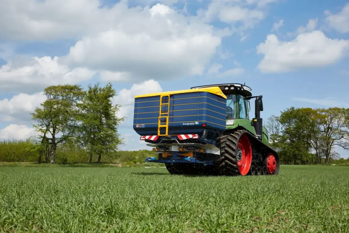 Zielony traktor z przyczepą rozsiewającą na polu. Na przyczepie widoczna nazwa "bogballe tabela wysiewu".