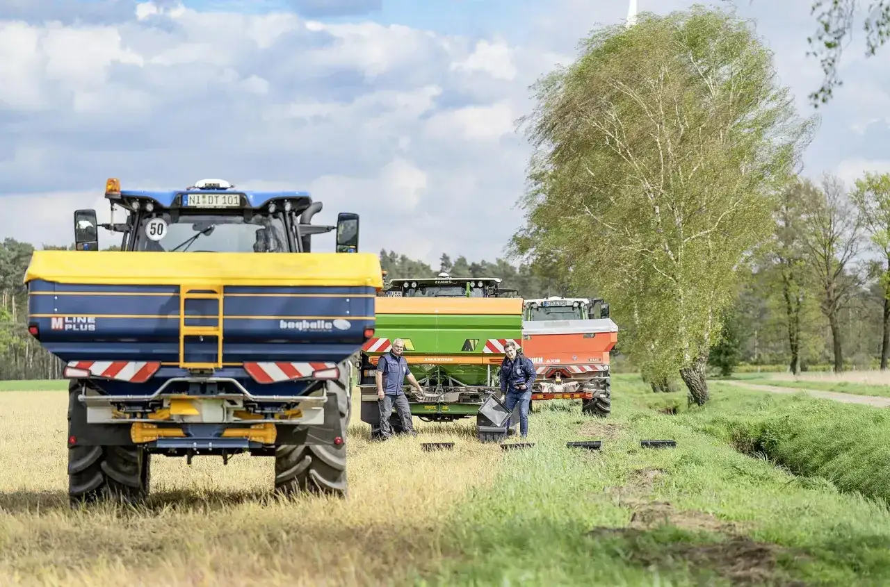 Rolnicy przygotowują rozsiewacze, w tym model Bogballe, do pracy. Na polu stoją trzy maszyny rolnicze, a dwaj mężczyźni sprawdzają sprzęt.